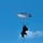 A person is suspended in mid-air beneath a small parachute or paraglider canopy against a clear blue sky. The individual appears to be in full gear, possibly military or tactical equipment, and is also connected to a vertical line or rope below them. The parachute canopy is partially deployed with a white and gray panel design.