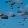 A large-scale military airborne operation showing numerous paratroopers descending from the sky with dark olive/grey parachutes deployed. The image captures what appears to be a mass tactical parachute jump, likely a military exercise or operation. Dozens of personnel are visible at various altitudes against a bright blue sky with scattered clouds.