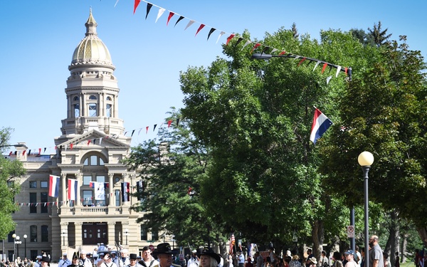 A patriotic parade is taking place on a tree-lined street in front of what appears to be the Wyoming State Capitol building with its distinctive gold dome. A white classic Ford Mustang convertible leads the procession, carrying passengers in naval and cowboy attire, followed by marchers holding a 'USS Constitution' banner. The event appears to be a summer celebration, likely related to Cheyenne Frontier Days or a similar civic occasion, with crowds lining the sidewalks.