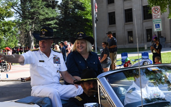 Vice Admiral Sean Buck and Mrs. Joanne Buck, identified as the 63rd Superintendent of the U.S. Naval Academy, are riding in a white convertible Ford Mustang during a parade. The Admiral is dressed in full white Navy dress uniform and both are wearing black cowboy hats, waving to the crowd. Spectators line the parade route in what appears to be a sunny outdoor civic event.