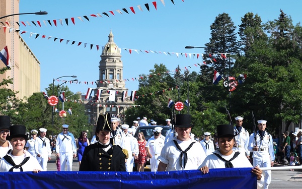 A parade is taking place on a sunny day along a decorated urban street, featuring personnel representing the USS Constitution, the oldest commissioned warship afloat, also known as 'Old Ironsides.' Participants are dressed in period naval uniforms and modern U.S. Navy whites, carrying a large blue banner identifying the vessel. A gold-domed government building is visible in the background, consistent with a state capitol.