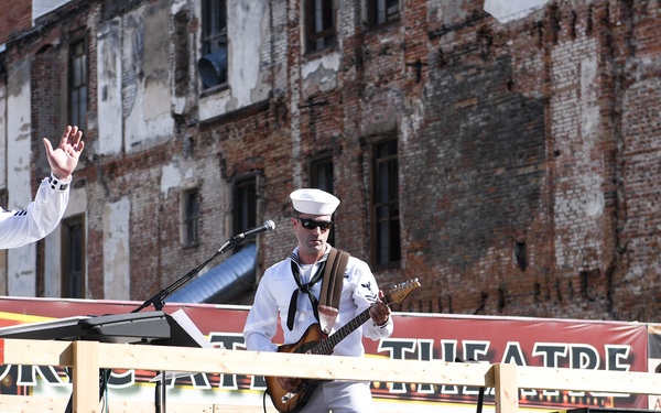 Three U.S. Navy sailors in white dress uniforms are performing on an outdoor stage mounted on a flatbed trailer. Two sailors are playing guitars while a third appears to be addressing or waving to the crowd. The performance is taking place in front of a weathered brick building with a partially visible 'Theatre' banner in the background.