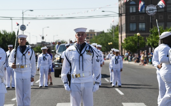 A group of U.S. Navy sailors in dress white uniforms are marching in a public parade along a city street. The formation includes personnel carrying rifles and wearing traditional Dixie cup caps with gold belt buckles. Spectators line the sidewalks in the background, and flags are visible on buildings along the parade route.