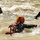 Two individuals are in turbulent, murky whitewater rapids. The foreground person wearing an orange helmet and a red vest labeled 'RESCUE' appears to be a water rescue professional actively swimming toward the background person. The second person, wearing a blue helmet, is being carried by the current and appears to be in distress or simulating a rescue scenario.