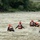 A group of water rescue personnel are wading through a fast-moving, turbid river. They are wearing matching red-and-black rescue vests labeled 'RESCUE' and orange safety helmets, suggesting a coordinated rescue operation or training exercise. The team is spread across the river in a deliberate formation, moving toward the far bank.