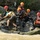 A group of water rescue personnel are conducting what appears to be a swift water rescue training exercise in a fast-moving, murky river. Multiple individuals wearing wetsuits, orange helmets, and life vests are assisting a civilian-dressed person onto a large rock mid-river. At least one person is visibly marked with 'RESCUE' on their vest, indicating professional emergency responders.