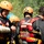A group of water rescue personnel are gathered together, wearing wetsuits, orange helmets, and red-and-yellow rescue life vests. One individual on the left has a bandaged/wrapped forearm and appears to be examining it. The team appears to be preparing for or debriefing after a water rescue operation.
