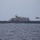 HSC 28 Helicopter Takes Off from the Flight Deck A U.S. Navy Littoral Combat Ship (LCS) is underway on open ocean under overcast skies. A military helicopter, consistent with an MH-60 Seahawk, is flying alongside the starboard side of the vessel. The scene appears to depict a naval aviation operation or flight operations exercise.