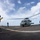 USS Sioux City and HSC 22 Sailors Participate in Flight Operations A military helicopter, appearing to be an MH-60 Seahawk, is on the flight deck of a naval vessel with its rotors spinning. Deck crew members in colored jerseys are positioned around the aircraft, performing flight operations. The scene takes place at sea under a partly cloudy sky, captured with a wide-angle fisheye lens.