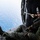 HSC 22 Sailor Participates in Flight Operations A military aircrew member is seated at the open door/ramp of a helicopter over open ocean water. The individual is wearing full flight gear including a flight helmet with visor, communications equipment, olive drab flight suit, tactical vest, and gloves. The perspective suggests the photo was taken from inside the aircraft looking toward the open rear or side hatch.