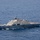 HSC 28 Helicopter Transfers Suspected Contraband An aerial photograph shows a U.S. Navy Littoral Combat Ship (LCS) underway on open blue ocean waters. The hull number '11' is visible on the bow, identifying it as USS Sioux City (LCS-11), a Freedom-class vessel. A military helicopter, appearing to be an MH-60 Seahawk, is conducting flight operations off the ship's starboard stern quarter with a load suspended on a hoist line.