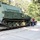 A large olive-drab military tracked armored vehicle is loaded on a flatbed lowboy trailer, parked on a concrete surface surrounded by trees. A single person wearing a hard hat and casual clothing stands beside the trailer, apparently overseeing or inspecting the transport. The vehicle appears to be an armored personnel carrier or similar tracked military platform in preserved or restored condition.