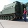 Three civilian workers are standing near a large military tracked armored vehicle, painted in olive drab green. The vehicle appears to be a heavy armored personnel carrier or similar military platform positioned on a concrete surface. The men appear to be inspecting or discussing the vehicle, possibly in a maintenance or delivery context.