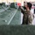 A U.S. Army soldier wearing a black face mask and MultiCam (OCP) uniform with a 3rd Infantry Division patch is inspecting or operating a component on a military armored vehicle painted in olive drab green. The setting appears to be an indoor maintenance facility or motor pool. Several other personnel, both military and civilian, are visible in the background.