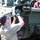 A man wearing gloves and glasses is working on the front section of a military tracked vehicle marked 'SPH0138', appearing to clean or apply material near the headlight assembly. A second person in a pink/magenta shirt is visible in the background on top of the vehicle. The vehicle is olive drab green with visible track wheels and tow chains.