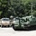 A military staging area shows a green self-propelled howitzer, likely an M109 Paladin, positioned in the foreground on a concrete pad. Behind it are several tan/desert-colored military trucks, including what appear to be Heavy Expanded Mobility Tactical Trucks (HEMTTs) or similar logistics vehicles. A civilian or soldier in casual clothing stands to the right observing the equipment.