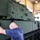 A bald civilian technician wearing work gloves and a blue polo shirt is inspecting or working on a large military armored vehicle painted in olive drab green, inside what appears to be a maintenance hangar or depot facility. The armored vehicle features a turret, track system, and bolt-on armor panels. A second individual in military uniform is visible in the background near the vehicle.