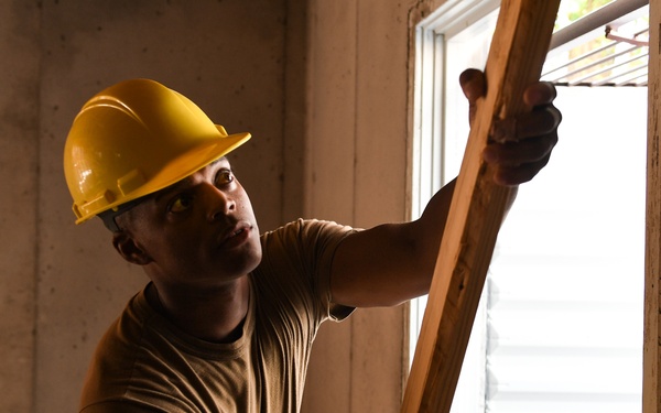 A worker wearing a yellow hard hat and military-style camouflage pants is handling a wooden beam or lumber near a window inside a concrete structure. The individual appears to be engaged in construction or renovation work, positioning or installing framing material. The setting suggests an active construction site with concrete block walls and a window opening.