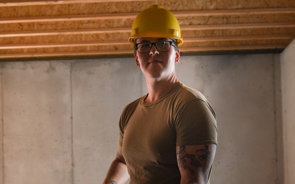 A construction worker wearing a yellow hard hat, glasses, and an olive-green t-shirt is carrying a large piece of drywall or building material inside an unfinished structure. The worker has visible tattoos on their right arm and is wearing work gloves. The setting appears to be an active construction site with exposed wooden framing, concrete walls, and a partially installed window.