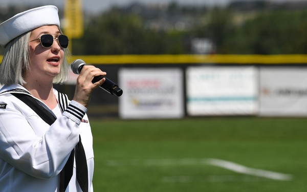 A person wearing a U.S. Navy dress white uniform and white sailor cap is singing into a handheld microphone on a baseball or sports field. The setting appears to be a pre-game ceremony, likely a national anthem performance. Sponsor banners and a green outfield fence are visible in the background.