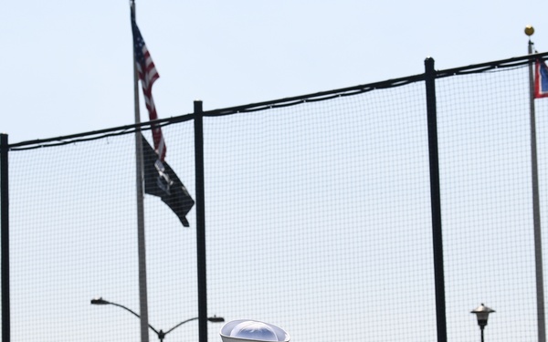 A person in a white U.S. Navy dress uniform stands at attention facing a large chain-link fence backdrop at what appears to be an outdoor stadium or athletic facility. Multiple flags are visible including the American flag, a POW/MIA flag, and what appears to be a state flag. Spectators can be seen seated in bleachers in the background.