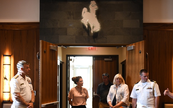 A group of military personnel and civilians are gathered in what appears to be a formal meeting or conference room with wood-paneled walls. The group includes Navy officers in white dress uniforms, an Army officer, an Air Force member in camouflage, and several civilians. They appear to be engaged in a briefing or tour of the facility.