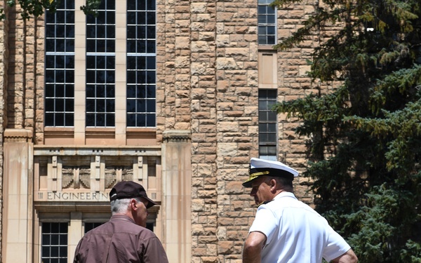 Two men are standing and conversing in front of a large stone building with 'Engineering' visible above the entrance. One man is dressed in a white naval officer uniform with a white peaked cap, while the other wears civilian clothing consisting of a brown shirt and khaki pants. The setting appears to be a university or institutional campus.