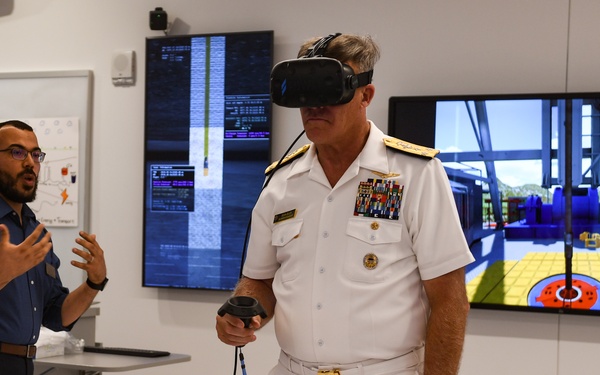A high-ranking U.S. Navy admiral in white dress uniform is using a virtual reality headset and hand controller in what appears to be a technology demonstration lab. A civilian presenter in a blue shirt stands nearby, gesturing and explaining the VR experience. Multiple display screens in the background show VR simulation content and code interfaces.