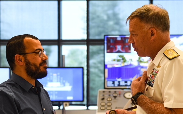 A U.S. Navy flag officer in white dress uniform is engaged in conversation with a civilian man in a blue shirt. The naval officer appears to be presenting or showing a challenge coin to the civilian. The setting appears to be a maritime simulation or operations training facility with multiple computer monitors and control panels visible.