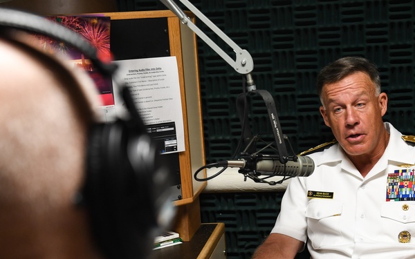 A U.S. Navy officer in white dress uniform is seated and speaking in what appears to be a radio broadcast studio. He is being interviewed by a person wearing headphones who is visible in the foreground out of focus. The setting includes acoustic foam panels on the walls and a professional microphone on a boom arm.