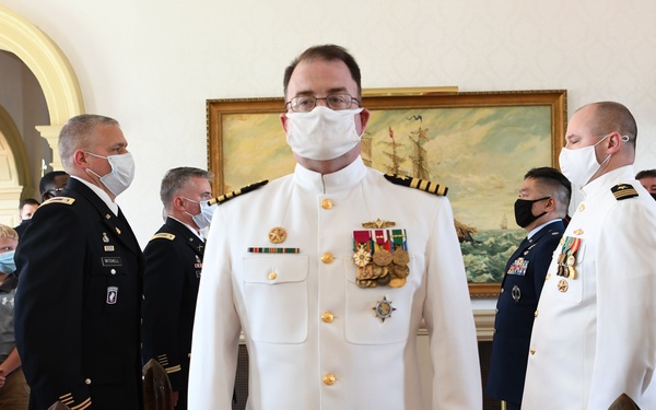 A formal military ceremony is taking place in an ornate indoor venue, featuring personnel from multiple branches of the U.S. Armed Forces wearing face masks. The central figure is a Navy officer in dress white uniform adorned with multiple medals and insignia, standing on a red carpet flanked by large brass shell casings. Personnel from the Army, Navy, and Air Force are visible, along with civilian attendees seated in the background.