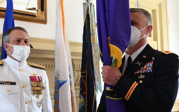 Two military officers in formal dress uniforms stand during what appears to be a change of command or official military ceremony. The officer on the left wears a white Navy dress uniform with multiple medals and decorations, while the officer on the right wears an Army dress blue uniform and holds a purple ceremonial flag. Both individuals are wearing white face masks, suggesting this event took place during the COVID-19 pandemic period.