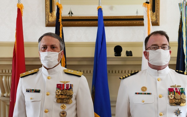 Two U.S. Navy officers in white dress uniforms stand side by side in a formal indoor setting, both wearing white face masks. The officer on the left displays a significantly larger number of medals and decorations, suggesting higher rank, while multiple military and organizational flags are arranged behind them. The setting appears to be an official ceremony or formal occasion.