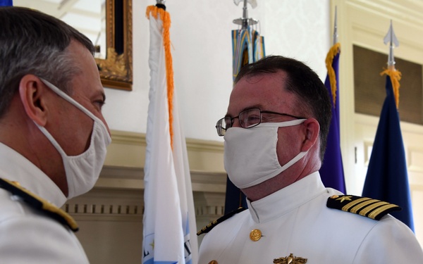 Two U.S. Navy officers in white dress uniforms are participating in a military decoration ceremony, with one officer pinning a medal onto the other's uniform. Both individuals are wearing white face masks, suggesting the event took place during the COVID-19 pandemic. The setting appears to be a formal military venue with flags and ornate architectural details visible in the background.