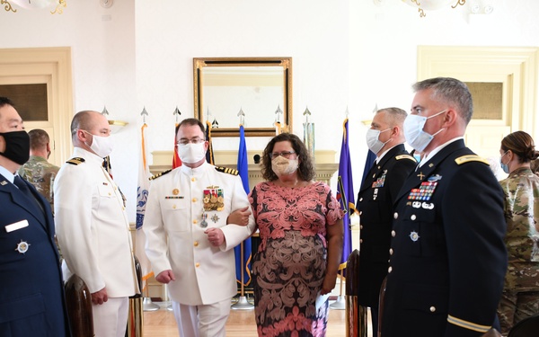 A formal military ceremony is taking place in an elegant indoor venue with a red carpet aisle. A naval officer in white dress uniform escorts a civilian woman in a floral dress down the aisle, flanked by military personnel from multiple branches including Army, Navy, and Air Force. All attendees are wearing face masks, consistent with COVID-19 era protocols.