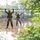 Two military personnel in full combat gear and camouflage uniforms are traversing a rope bridge obstacle over a murky brown body of water. They are gripping upper ropes with both hands while balancing their feet on lower ropes, suggesting a military training exercise. The setting is a wooded, humid environment with lush green foliage surrounding the water obstacle.