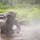 A soldier in full combat gear is crawling through a muddy water obstacle, heavily covered in mud and water. The individual is wearing a helmet, protective goggles, and tactical gloves while holding what appears to be a rifle. The scene appears to be a military training exercise, likely an obstacle course, with smoke or mist visible in the foreground.