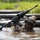 A soldier in full combat gear is wading through a muddy water obstacle, holding a rifle elevated above the water to keep it dry. The soldier is wearing camouflage uniform, tactical helmet, and black gloves, and appears to be navigating a military training course. Large black cylindrical obstacles or logs are visible in the background near the water.