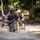 Multiple soldiers in camouflage uniforms and helmets are running through a shallow muddy waterway while carrying rifles. Their uniforms and gear are visibly soaked and mud-covered, suggesting an ongoing field training exercise. The wooded background and controlled environment indicate a military training course rather than a combat scenario.