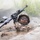 A soldier in full camouflage gear is navigating through a muddy water obstacle, holding a rifle above their head to keep it dry. The individual appears to be participating in a military training exercise, as indicated by the controlled environment with barbed wire and smoke effects. The soldier is fully submerged to chest level while maintaining weapon discipline.