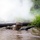 A soldier in full combat gear is low-crawling through a muddy water obstacle during what appears to be a military training exercise. The soldier is holding an assault rifle above the water while navigating under barbed wire and a log barrier. White smoke is visible in the background, consistent with a simulated combat training environment.