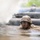 A soldier in full combat gear, including a helmet and protective goggles, is crawling through a muddy water obstacle course with barbed wire overhead and smoke present. The individual is completely covered in mud with only their head visible above the murky water. This appears to be a military training exercise, likely a low-crawl obstacle course under simulated combat conditions.