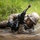 A soldier in digital camouflage uniform is crawling through a muddy water obstacle during what appears to be a military training exercise. The individual is holding a rifle elevated above the water while navigating under barbed wire. Face paint and full combat gear suggest this is a structured field training or obstacle course exercise.