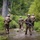 Four soldiers in camouflage uniforms are wading through a shallow muddy waterway in a forested environment. Their uniforms and gear are heavily soiled with mud, suggesting an extended field exercise or training operation. One soldier faces the camera while carrying a rifle, while the others move forward through the water.