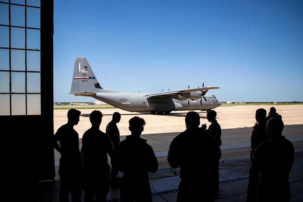 A group of silhouetted personnel stand inside a hangar observing a U.S. Air Force C-130 Hercules transport aircraft on the tarmac. The aircraft bears the tail markings 'AMC' and serial number '83172', with 'U.S. Air Force' visible on the fuselage. The scene appears to be a briefing, observation, or arrival event at a military airfield under clear blue skies.