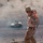 A military personnel in desert camouflage (MARPAT pattern) stands on an elevated rocky ridge overlooking a flat desert terrain. The soldier is equipped with an orange-marked training vest/harness and a backpack, holding what appears to be a small device or card. Below on the desert floor, a tan HMMWV (Humvee) military vehicle is visible with personnel in the turret, surrounded by dust.