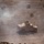 An aerial/elevated view of a tan-colored military HMMWV (Humvee) operating in an arid desert environment. A gunner is visible in the turret position, and smoke or dust is present to the left of the vehicle. The terrain is dry, rocky, and sparsely vegetated.