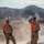 Two military personnel in desert camouflage uniforms and bright orange safety vests stand on elevated ground observing an active firing range in an arid desert environment. Smoke plumes and what appears to be an illumination or pyrotechnic round are visible downrange against a backdrop of rocky mountains. The orange vests are marked with identification numbers '43B' and '44H', indicating range safety officer or observer roles.