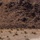 A single military HMMWV (Humvee) traverses a flat, arid desert terrain, kicking up a dust trail behind it. The vehicle is moving across what appears to be a dry lakebed or desert floor with sparse scrub vegetation. A rocky, barren hillside forms the backdrop in the upper portion of the image.