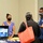 Three individuals are gathered at an informational booth or table at what appears to be a community event or job fair. Two booth representatives wearing face masks are engaging with a visitor carrying a red tote bag. A monitor on the table displays a slideshow with slides labeled 'Community Involvement' and 'Care &amp; Support.'